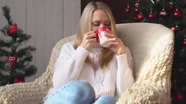 Una donna su una sedia che beve caffè o tè su uno sfondo di albero di Natale . — Video Stock