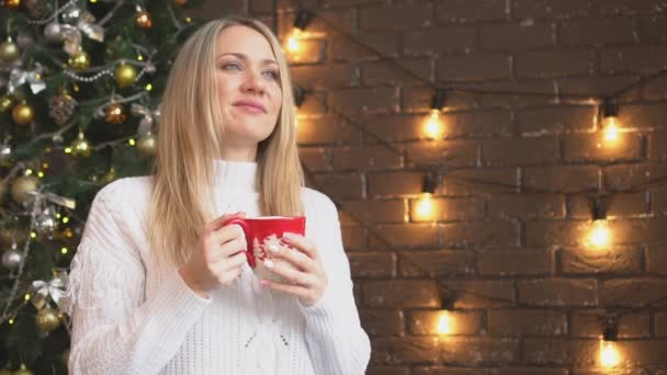 Jul, en ung kvinna i en vit tröja som dricker kaffe eller te från en mugg. — Stockvideo