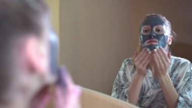 Bir kadın bir ayna önünde yüz krem maske koyar.
