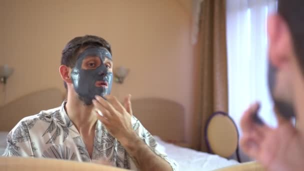 Un uomo davanti a uno specchio sul volto di una maschera cosmetica di argilla . — Video Stock