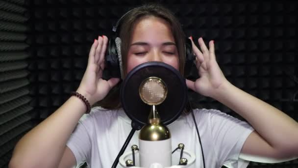 Uma jovem mulher em fones de ouvido cantando no microfone no estúdio de gravação . — Vídeo de Stock