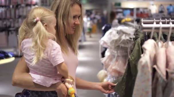 Uma mulher com um filho a comprar roupas numa loja infantil. Mãe e filha no shopping . — Vídeo de Stock