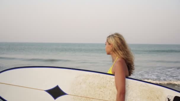 Блондинка-серфингистка прогуливается по пляжу с доской для серфинга — стоковое видео