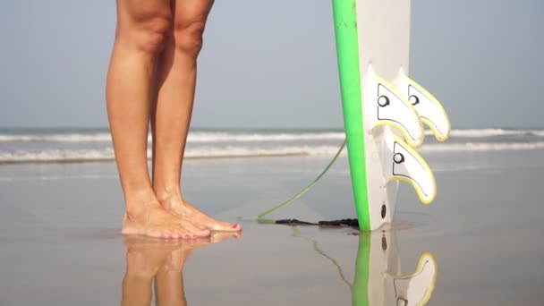 En flicka står på sanden mot bakgrunden av havet med en surfbräda — Stockvideo