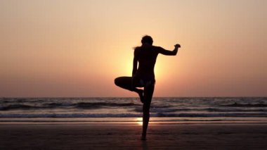 Gün batımında plajda yoga yapan genç bir kadın.