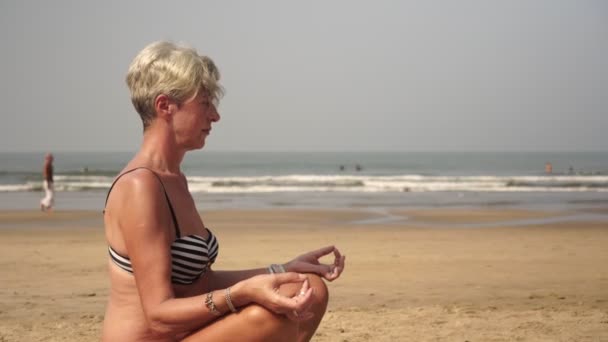 Mujer madura jubilada haciendo yoga al aire libre. La abuela está meditando en la playa — Vídeos de Stock