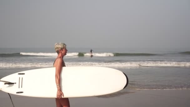 Dojrzała kobieta seniorek spaceruje po plaży z deską surfingową. — Wideo stockowe