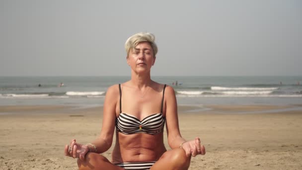 Idős nő idős állampolgár meditál a strandon lótuszpozícióban. — Stock videók