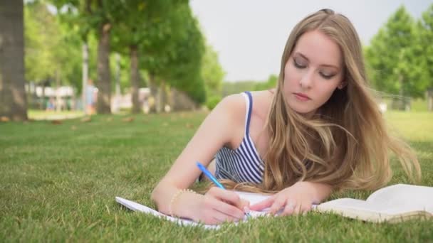 Uma jovem estudante em um parque jaz em um gramado escreve em um caderno . — Vídeo de Stock