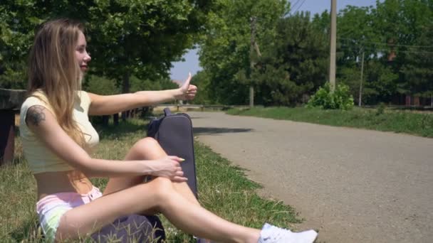 A young woman hitchhikes and sits with a suitcase on the side of the road — Stock Video