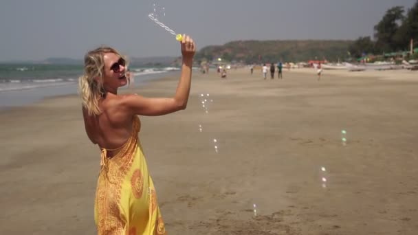 Jonge vrouw in een zomer jurk is blazen bubbels op het strand, slow motion — Stockvideo