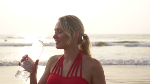 Žena pije vodu z láhve na pozadí západu slunce nad mořem — Stock video