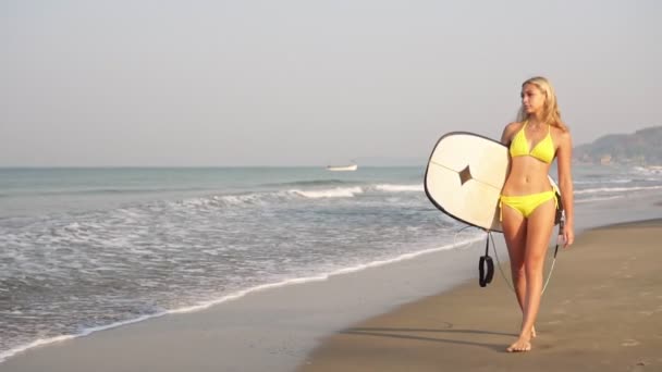 아름다운 한 젊은 여자가 파도타기 판을 들고 해변을 따라 걷는다. 느린 동작 — 비디오