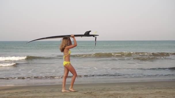 Молодая женщина прогуливается по морскому пляжу с доской для серфинга, замедленным движением — стоковое видео