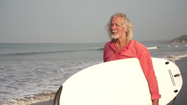 El viejo jubilado a la edad va a practicar deportes. Robusto abuelo de pelo gris en la playa con una tabla de surf. Concepto de estilo de vida activo de jubilación — Vídeos de Stock