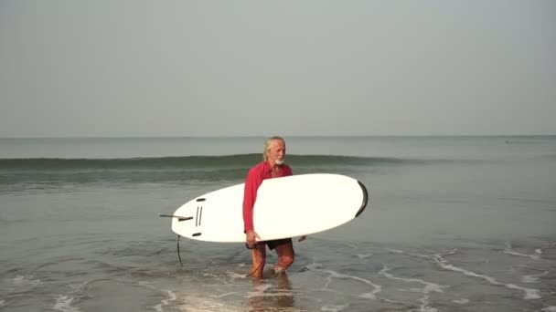 Старий виходить з моря з дошкою для серфінгу. Старший пенсіонер займається спортом. Літня людина збагачує і веде активний спортивний спосіб життя — стокове відео