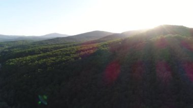 Hava manzarası: Ormanın üzerinde gün batımı ya da gün doğumu. Güneş ışınları, dağlar ve yeşil ağaçlı tepeler