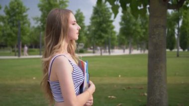 Genç mutlu kız öğrenci elinde defter ve ders kitabıyla parkta yürüyor.