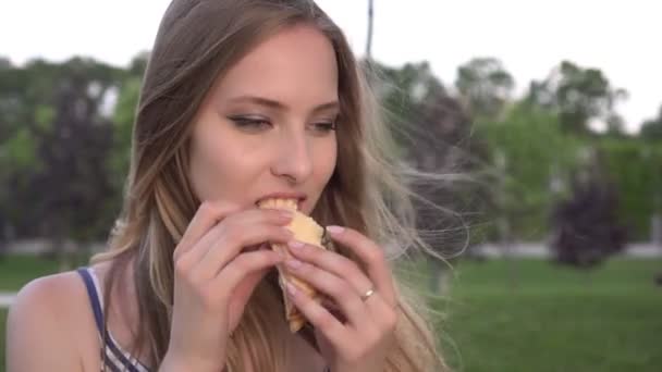Young attractive woman eating a sandwich outdoors. Portrait of a woman with fast food — Stock Video