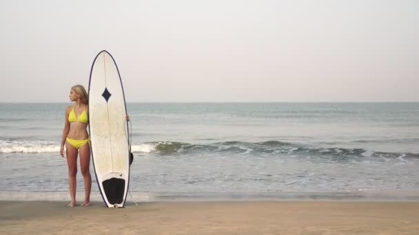 Jonge aantrekkelijke vrouw surfer in een gele bikini met een surfplank op de achtergrond van de zee — Stockvideo