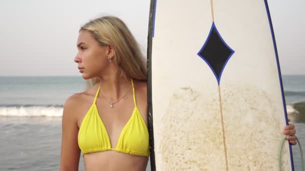 Portret van een jonge aantrekkelijke vrouwelijke surfer in een geel zwempak met een surfplank op de achtergrond van de zee — Stockvideo