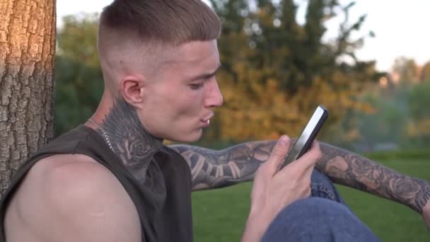 Молодий сучасний чоловік з татуюваннями використовує смартфон на відкритому повітрі в парку — стокове відео
