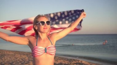Amerikan bayrağı taşıyan çekici genç bir kadın kumlu sahil boyunca yürüyor.