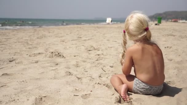 Niña solitaria sentada en la playa del mar en la arena. El niño juega en la playa contra el fondo del mar — Vídeos de Stock