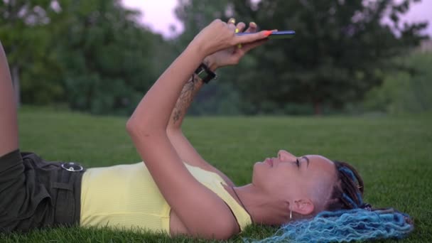 Wanita muda modern menarik terletak di rumput di taman dan menggunakan smartphone — Stok Video