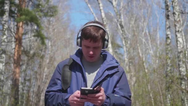Ένας νεαρός άντρας βόλτες στο πάρκο, να ακούτε μουσική μέσω των ακουστικών. Πρωινή βόλτα στο φρέσκο αέρα άνοιξη πάρκο. — Αρχείο Βίντεο