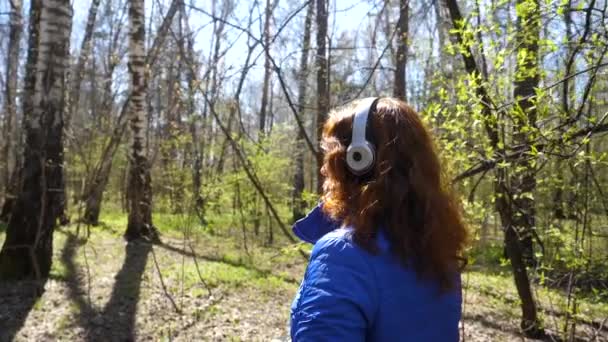 Ein junges Mädchen im Park, das über Kopfhörer Musik hört. eine Frau spaziert durch den Frühlingspark und genießt die Musik. — Stockvideo