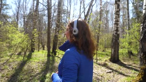 ヘッドフォンで音楽を聴く公園の少女。女性は、春の公園を歩くし、音楽を楽しむ. — ストック動画