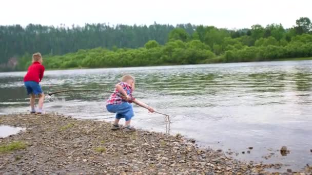 Dos niños juegan en la orilla del río. Tira piedras, haz salpicaduras de agua. Hermoso paisaje de verano . — Vídeo de stock