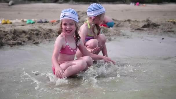 Dos chicas salpicando agua en la playa. Gemelos vierten agua en un día caluroso de verano, la risa y el buen humor de los niños — Vídeos de Stock