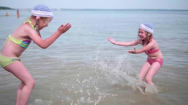 Две девушки брызгают водой на пляж. Близнецы наливают воду в жаркий летний день, смех и хорошее настроение детей — стоковое видео