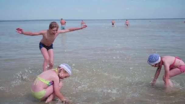 Две девушки и парень, брызгающий водой на пляж. Близнецы наливают воду в жаркий летний день, смех и хорошее настроение детей. Летний отдых на море . — стоковое видео