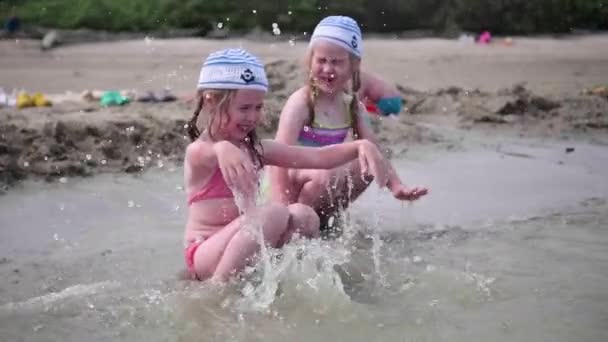 Két lány és fröccsenő víz a strandon. Ikrek vizet öntsünk egy forró nyári napon, a nevetés és a jó hangulat, a gyermekek — Stock videók