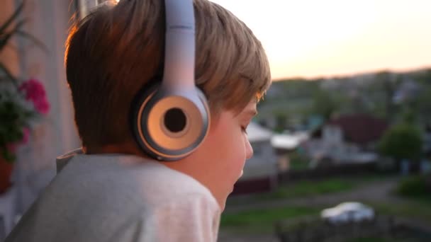 Um jovem fica perto da janela e coloca fones de ouvido para ouvir música. Fundo borrado com pôr do sol, adolescente desfrutando de música em fones de ouvido . — Vídeo de Stock