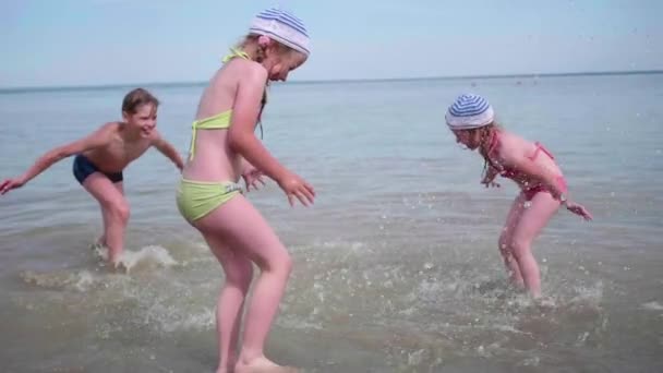 Dos chicas y un tipo salpicando agua en la playa. Los gemelos vierten el agua en el día caluroso veraniego, la risa y el humor bueno de los niños. Vacaciones de verano en el mar . — Vídeo de stock