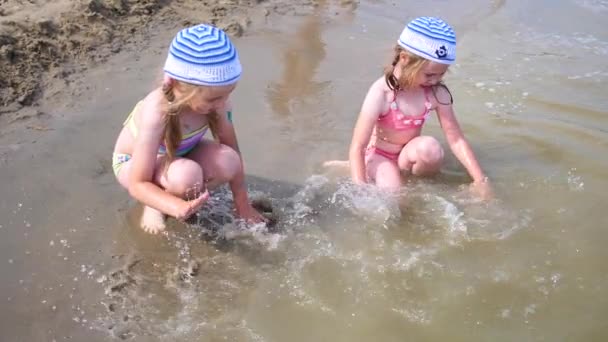 Twee meisjes spatten op het strand. Tweeling giet water op een warme zomerdag, een lach en een goed humeur van kinderen — Stockvideo