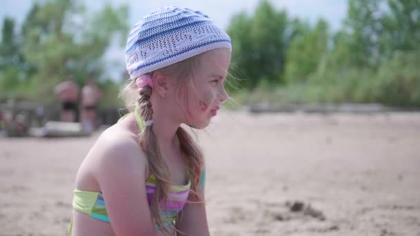 Little girl sitting on the beach, hot summer day. Carefully and thoughtfully looking into the distance. — Stock Video