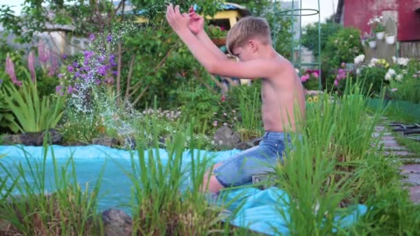 Pojken sitter vid en liten sjö, runt sjön växer blommor och gräs. Barnet skapar stänk av vatten med fötterna. Varm sommardag. Lycklig barndom — Stockvideo