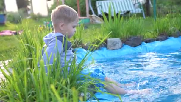 O menino está sentado em um pequeno lago, ao redor do lago crescem flores e grama. A criança cria salpicos de água com os pés. Dia quente de verão. Infância feliz — Vídeo de Stock