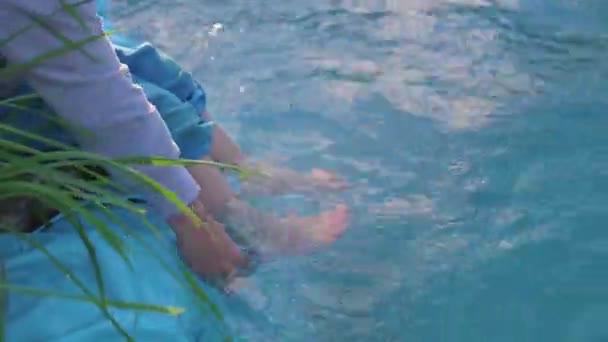 Pojken sitter på en liten sjö. Barnet skapar stänk av vatten med fötterna. Varm sommardag. Fötter närbild — Stockvideo