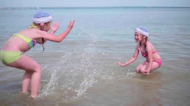 Две забавные девчонки плеснули водой на пляж. Близнецы наливают воду в жаркий летний день, смех и хорошее настроение детей — стоковое видео