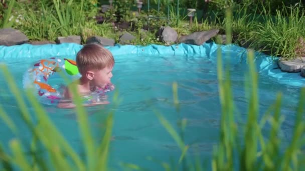 El niño nada en un círculo inflable en un pequeño estanque. Área de jardín, flores y plantas alrededor del lago. Feliz infancia. — Vídeo de stock