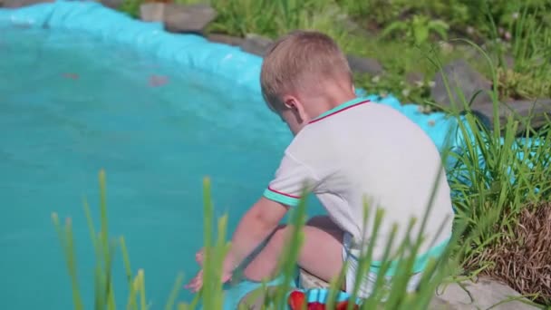 The boy is sitting at a small lake. The child creates splashes of water with his feet. Hot summer day. Happy childhood — Stock Video
