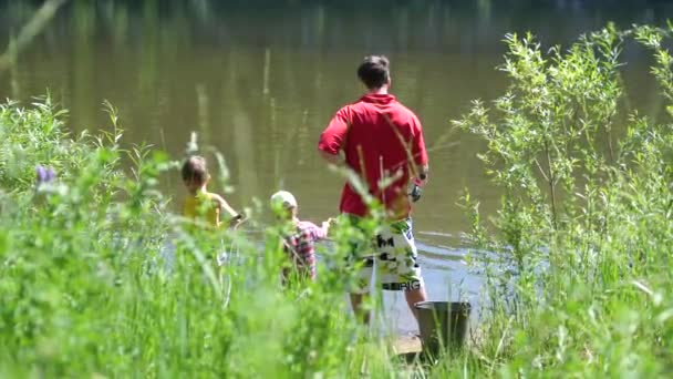 Dos niños pequeños y un joven padre pescando en la orilla del río. Hermoso paisaje de verano. recreación al aire libre . — Vídeo de stock