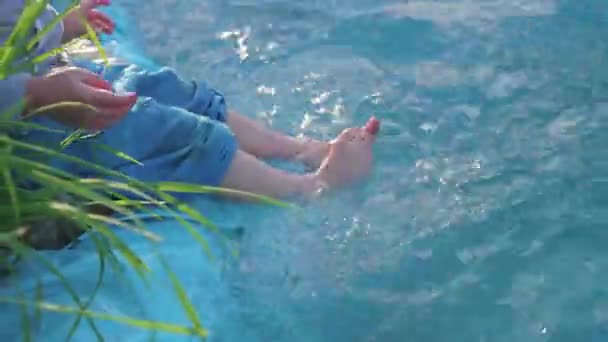那个男孩正坐在一个小湖边。这孩子用脚制造水花。炎热的夏日。快乐童年 — 图库视频影像