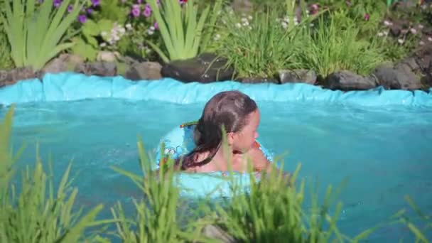 Κορίτσι κολύμβησης σε μια μικρή λιμνούλα. Το παιδί βρίσκεται σε δροσερό νερό σε μια καυτή καλοκαιρινή μέρα. Ευτυχισμένη παιδική ηλικία. Λουλούδια και γρασίδι αναπτύσσεται γύρω από τη λίμνη — Αρχείο Βίντεο